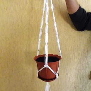 以下が含まれることがあります： 白いマクラメのプラントハンガーと茶色の鉢。ハンガーはコットンコードで作られており、下部にタッセルが付いています。