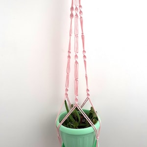 May include: A pink macrame plant hanger with a light green pot and a small plant inside.