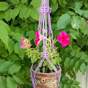 May include: A purple macrame plant hanger with a terracotta pot and pink flowers. The hanger is made with a knot pattern and has a decorative design.