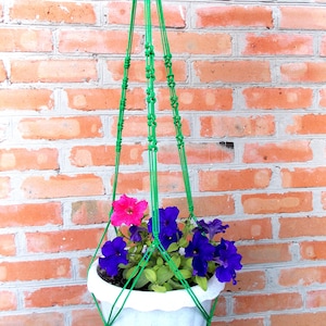 May include: A white hanging planter with purple and pink flowers, suspended from a green macrame cord.