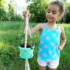 Puede incluir: Una persona joven sosteniendo un macramé para colgar plantas con una maceta azul claro y una planta verde. El macramé está hecho de cordón de algodón blanco. La persona lleva una camiseta sin mangas azul con flamencos rosas y pantalones cortos blancos.