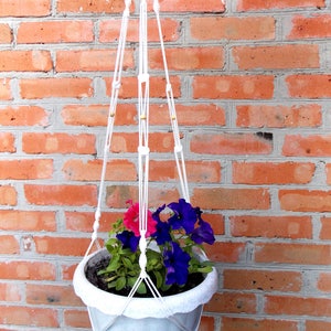 May include: White macrame plant hanger with a white ceramic pot and pink and purple flowers hanging against a red brick wall.