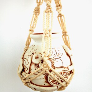 May include: A white ceramic planter with a brown floral design is suspended from a macrame hanger made of cream-coloured rope.