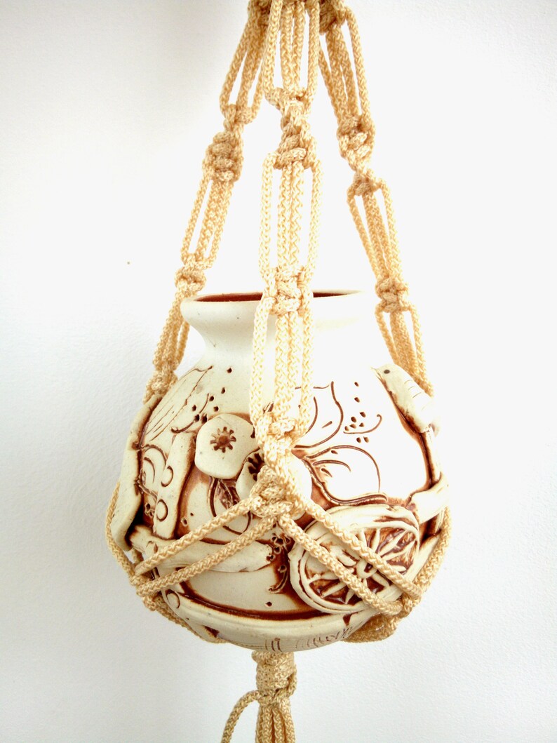 May include: A white ceramic planter with a brown floral design is suspended from a macrame hanger made of cream-coloured rope.
