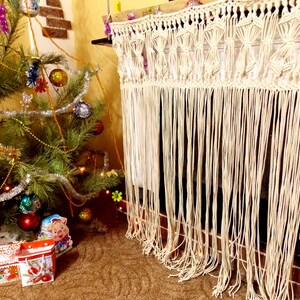 Könnte beinhalten: Ein weißer Makramee-Kaminsims-Schal mit Fransen. Der Schal hängt über einem gemauerten Kamin und im Vordergrund steht ein Weihnachtsbaum.