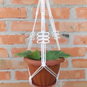 Puede incluir: Un macetero de macramé blanco con una maceta marrón y una planta verde en su interior. El macetero está colgado de una pared de ladrillo.