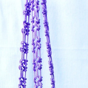May include: A close-up of a purple macrame plant hanger with a knotted design. The hanger is made of multiple strands of cord and is hanging against a white background.