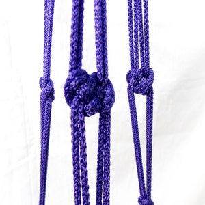 May include: Close-up of a purple macrame plant hanger. The hanger features intricate knots and braided cords, creating a decorative and functional design. The vibrant purple color adds a pop of color against the white background. Perfect for displaying plants.