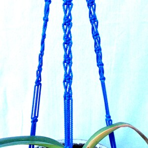 May include: A blue macrame plant hanger with three strands. The hanger is made of thick cotton cord and has a knot pattern. The hanger is holding a potted plant.