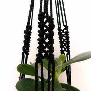 Puede incluir: Un colgador de plantas de macramé negro con intrincados detalles de nudos. El colgador de plantas está suspendido, mostrando su diseño tejido. Se ven hojas verdes de plantas dentro del colgador, creando un contraste. Adecuado para uso en interiores o exteriores.