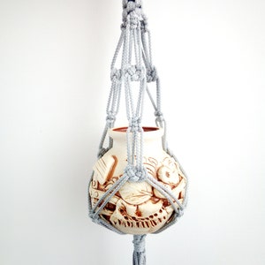 Puede incluir: Un macetero de macramé gris con una maceta de cerámica blanca con un diseño floral marrón. La maceta cuelga del macetero de macramé.