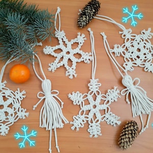 May include: Six white macrame snowflake ornaments with tassels hanging on a wooden surface. The ornaments are decorated with white string and have a knot design. There are also pine cones and a tangerine on the surface.