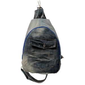 Op de afbeelding: Een druppelvormige sling-rugzak gemaakt van licht- en donkerblauwe spijkerstof. De tas is voorzien van een voorvak met klep, een opvallende blauwe ritssluiting en een enkele verstelbare riem die aan de boven- en onderkant is bevestigd.
