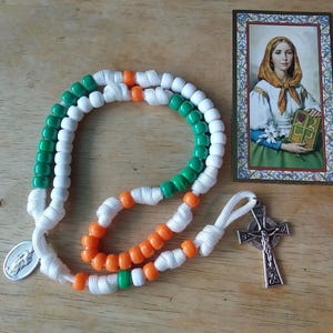 May include: A rosary with green, white, and orange beads, a silver crucifix, and a small picture of a saint. The beads are strung on white cord. The image of the saint is in a decorative frame.