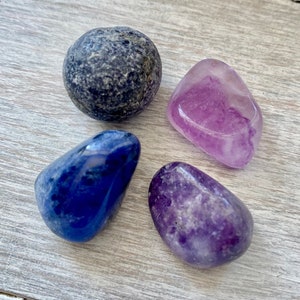 May include: Four tumbled stones, one dark blue, one light blue, and two shades of purple. The stones are smooth and polished.
