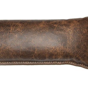 May include: A long, brown leather cushion with a distressed finish. The cushion has a small leather tag attached to it with the text "The Original" printed on it.