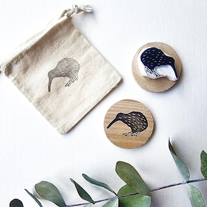 May include: A set of kiwi bird-themed items. Includes two wooden coasters with a kiwi bird design, a small drawstring bag with a kiwi bird print, and a sprig of green leaves. The kiwi bird design is black and white.