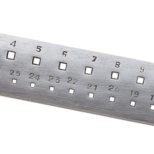 Può includere: Un calibro metallico rettangolare argentato con fori quadrati e numeri da 1 a 30. Il calibro ha una texture in metallo spazzolato. I fori quadrati sono distanziati uniformemente e diminuiscono di dimensione.