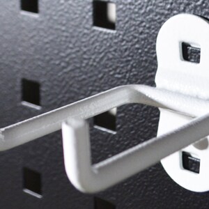 May include: A white metal pegboard hook with a curved design. The hook is mounted on a black pegboard with square holes. The hook has a textured surface and is designed to hold items.
