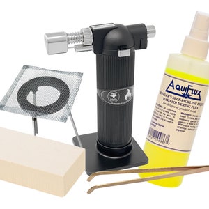 May include: A jewelry making kit with a black torch, a yellow bottle of Aquiflux Jeweler's Self-Pickling Liquid Hard Soldering Flux, a white fire brick, a metal mesh ring stand, and a pair of brass tweezers.