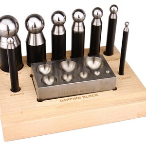 Puede incluir: Un juego de 8 herramientas de dapping de metal y un bloque de dapping con 8 depresiones de diferentes tamaños. Las herramientas están dispuestas en un soporte de madera con medidas etiquetadas en milímetros.