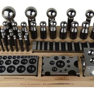 Op de afbeelding: Een klassieke dappingset met een houten basis en een verscheidenheid aan metalen dappinggereedschappen en -blokken. De set bevat een dappingplaat met 24 gaten, een dappingblok en een verscheidenheid aan dappingponsen.