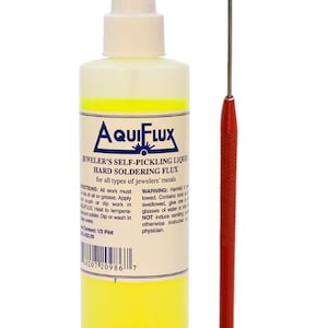 Könnte beinhalten: Eine Flasche AQUIFLUX selbstbeizende Flüssigkeit Hartlötflussmittel für Juweliere mit einem rotgriffigen Lötdorn. Die Flasche ist überwiegend weiß mit gelbem Boden.