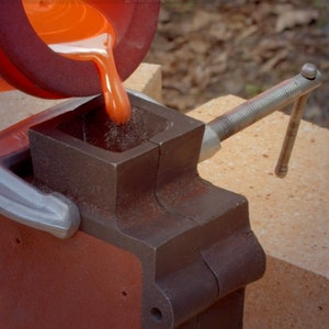May include: Molten orange liquid being poured into a metal mold clamped to a brick surface.