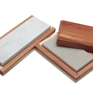 Op de afbeelding: Twee rechthoekige slijpstenen in een houten doos. De stenen zijn wit en grijs, en de doos is gemaakt van lichtbruin hout.