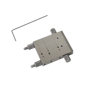 May include: A silver metal tool with adjustable knobs and a small, bent metal rod. The tool features multiple slots and holes, and the knobs are on the sides. The rod is positioned near the tool.