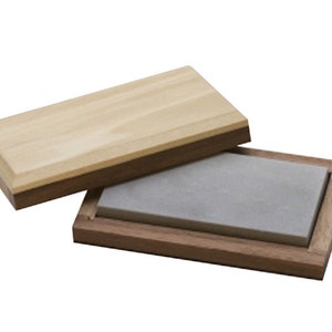 Könnte beinhalten: Ein grauer Schleifstein in einer Holzkiste mit einem hellen Holzdeckel. Die Kiste ist aus dunklem Holz und hat einen hellen Holzrahmen.