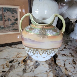 May include: A white porcelain vase with a gold and green floral design. The vase has a wide mouth and two handles. The vase is sitting on a granite countertop.