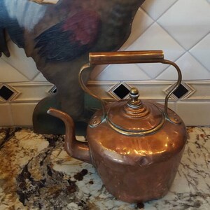 May include: A copper teapot with a wooden handle and a brass spout. The teapot is sitting on a countertop with a painted rooster figurine in the background.