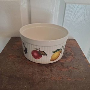 May include: White ceramic baking dish with a fruit design. The dish has a red apple, a yellow lemon, and purple grapes on it.
