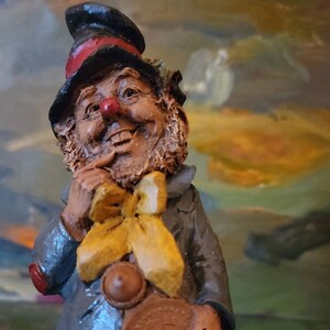 May include: A whimsical figurine of a smiling leprechaun, wearing a black hat with a red band, a gray coat, and a yellow bow tie. The leprechaun holds a large brown coin. The figurine has a red nose and a detailed, textured appearance.