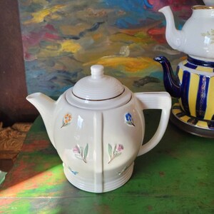 May include: A white ceramic teapot with a floral design. The teapot has a lid and a handle. The teapot is sitting on a green table.