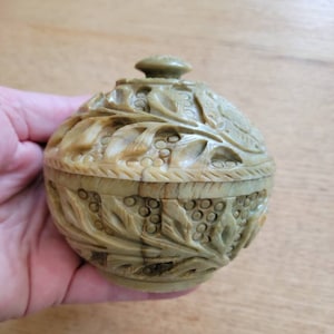 May include: A round, green stone trinket box with a carved floral design. The lid is closed and the box is sitting in a hand.