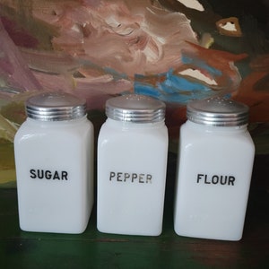 May include: Three white ceramic canisters with silver lids. The canisters are labeled "Sugar", "Pepper", and "Flour".