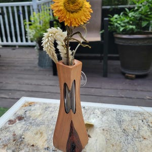 May include: A wooden vase with a clear glass insert, holding a dried sunflower and other dried flowers. The vase has a unique, curved design and is made of light brown wood with dark brown accents.