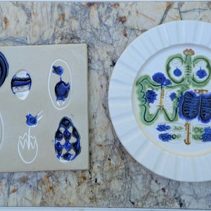 May include: A white ceramic plate with a blue and green design. The plate has a scalloped edge and is decorated with a stylized floral pattern. There is also a smaller ceramic tile with a blue and white design.