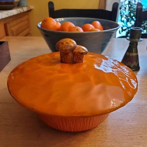 May include: Orange ceramic casserole dish with a lid featuring two mushroom-shaped handles. The dish has a textured, rippled surface.