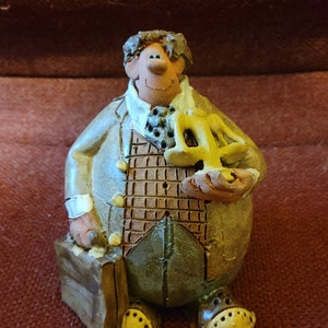 May include: A ceramic figurine of a man wearing a brown and blue suit, holding a scale of justice in his right hand and a brown briefcase in his left hand. The figurine has a smiling face and is painted in a rustic style.