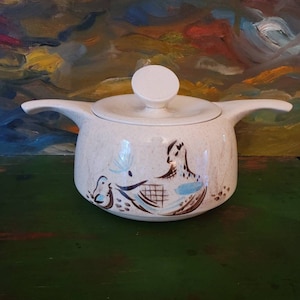 May include: A white ceramic sugar bowl with a lid and a brown and blue bird design. The bowl has a speckled glaze and a round shape.
