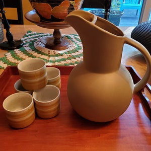 May include: A set of four small ceramic cups with brown and white stripes, stacked on a wooden tray. A large, light brown ceramic pitcher sits next to the cups.