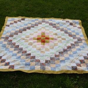 May include: A patchwork quilt with a diamond pattern in the center. The quilt is made of various fabrics in shades of blue, white, pink, and yellow. The center diamond is made of red, orange, and yellow fabrics.
