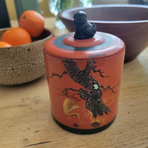 May include: A red and black cylindrical container with a black animal figure on the lid. The container has a black and gold design with a tree branch and birds.