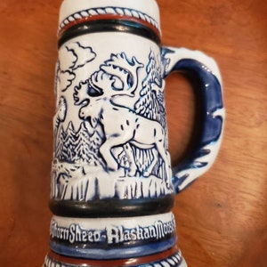 Pode incluir: Caneca de cerveja de cer&acirc;mica branca com um design de alce azul e branco. A caneca tem um design de corda marrom ao redor da parte superior e inferior. O texto "Horn Sheed - Alaskan Moose" est&aacute; impresso na parte inferior da caneca.