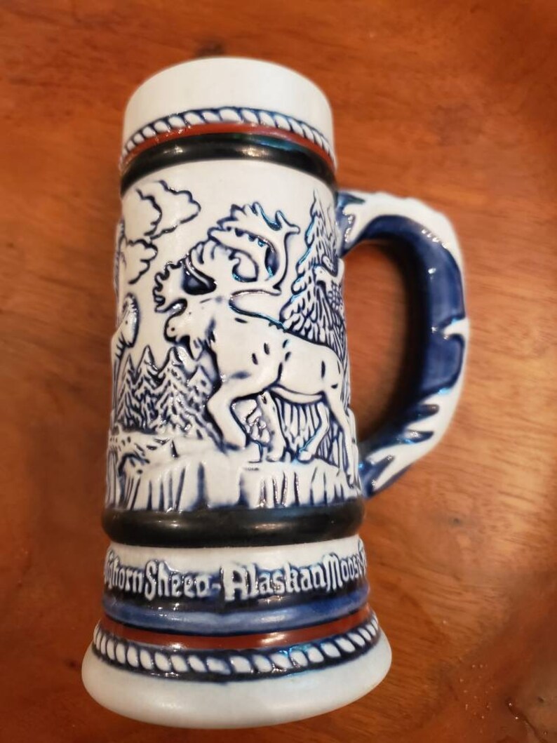 Pode incluir: Caneca de cerveja de cer&acirc;mica branca com um design de alce azul e branco. A caneca tem um design de corda marrom ao redor da parte superior e inferior. O texto "Horn Sheed - Alaskan Moose" est&aacute; impresso na parte inferior da caneca.