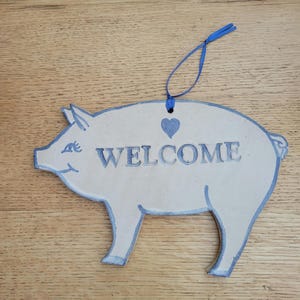 May include: A white ceramic pig shaped welcome sign with blue trim and a blue heart. The sign says "WELCOME" in blue lettering.