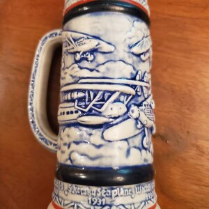 Pode incluir: Uma caneca de cerveja de cer&acirc;mica branca com detalhes em azul e vermelho. A caneca apresenta um design em relevo de hidroavi&otilde;es antigos voando nas nuvens. O texto "Sikorsky Seaplane Waterbury 1931" est&aacute; impresso na parte inferior da caneca.